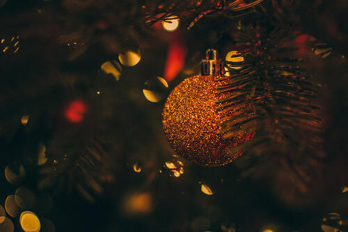 Bilde av ei gullfarga julekule blant edelgrangreiner og antydning til juletrelys og noko raudt i bakgrunnen.